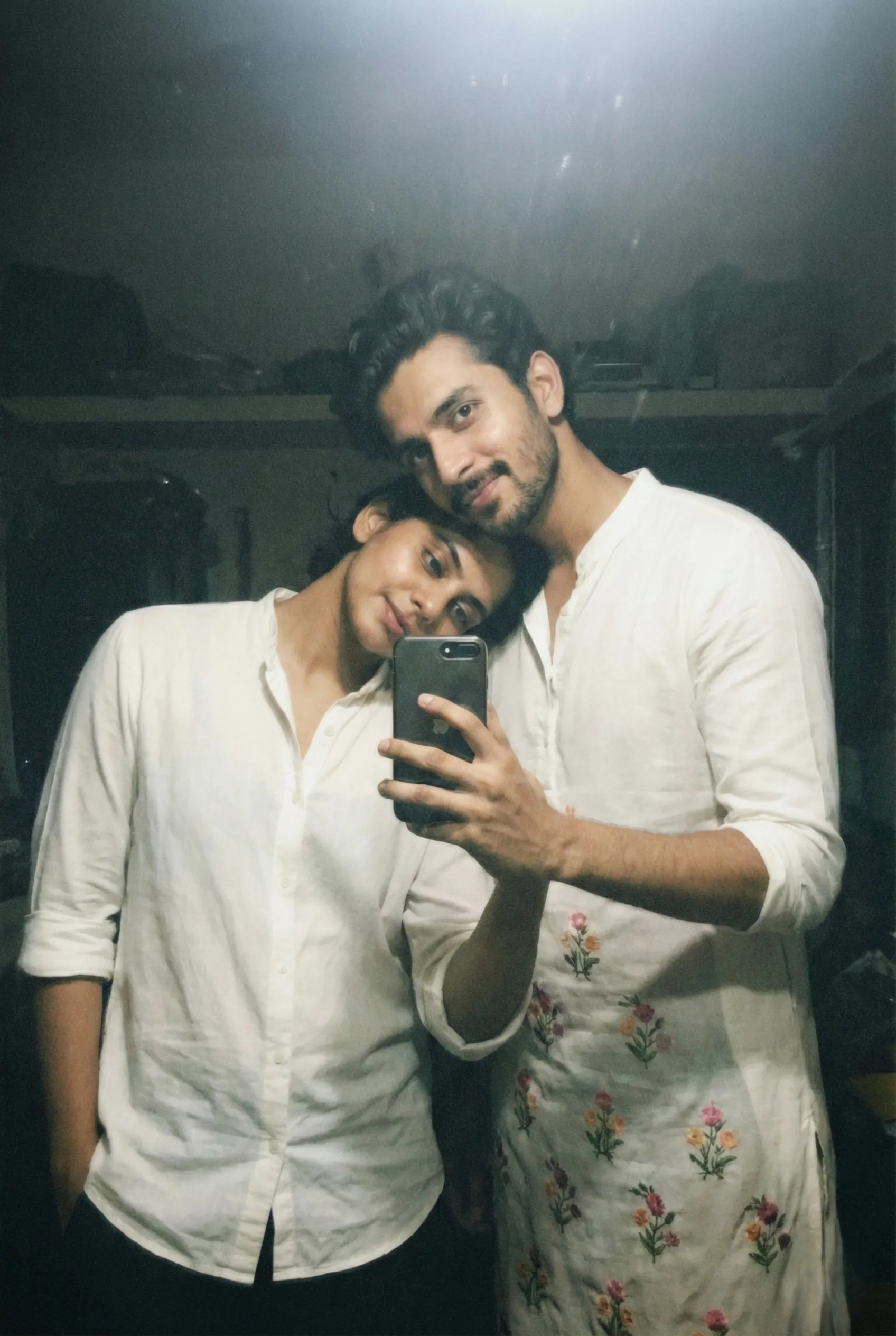 Grainy lo-fi mirror selfie of a couple; woman in a white shirt holding an iPhone, man in a floral embroidered kurta leaning his head on her, soft flash photography aesthetic.